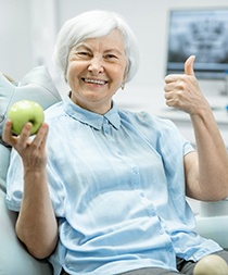 An older woman holding an apple and giving a thumbs-up after receiving her new dental implants
