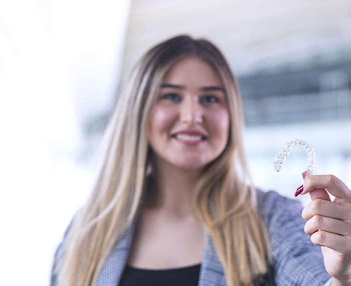 A young woman wearing a jacket and holding an Invisalign aligner in her left hand while smiling